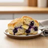 A close-up of a warm, golden-brown easy blueberry biscuits split in half, revealing flaky layers and juicy blueberries, with melting butter inside, on a rustic plate