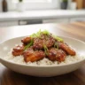 A macro close-up of Food Court Style Glazed Chicken over white rice, garnished with green onions and sesame seeds, perfectly centered in a bowl on a rustic wooden counter. The chicken glistens with a sticky, dark amber glaze. This is the ultimate food court glazed chicken