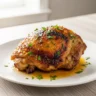 A close-up of a perfectly roasted Golden Lemon Honey Glazed Chicken thigh, garnished with fresh parsley, plated on a white dish, showcasing a delicious lemon honey chicken recipe