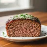 A macro close-up of a perfectly sliced Ultimate Garlic Parmesan Meatloaf on a white plate, garnished with fresh parsley and Parmesan, showing its juicy interior and tangy-sweet glaze, perfect for a comforting garlic parmesan meatloaf recipe