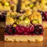 Macro close-up of a perfectly cut Luscious Blackberry Pistachio Dream Bar, showcasing its layered textures: golden pistachio crumble, vibrant purple blackberry filling, and buttery shortbread crust.