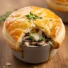 Macro close-up of a golden, flaky Rustic Gruyère & Mushroom Mini Pot Pie in a ramekin, garnished with fresh thyme.