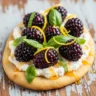Close-up of a slice of Berry Bliss Ricotta Flatbread with fresh blackberries, basil, lemon zest, and honey drizzle on a rustic wooden board.