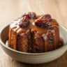 A close-up of a warm, date-studded sticky toffee pudding drenched in glossy, buttery toffee sauce in a rustic bowl.
