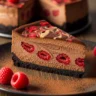 Macro close-up of a slice of Ultimate Dark Chocolate Raspberry Cheesecake with vibrant red swirls, rich dark chocolate, and a crisp chocolate cookie crust, garnished with fresh raspberries.