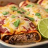 Macro close-up of a freshly baked black bean enchilada with melted cheese, red sauce, cilantro, and lime on a rustic wooden table.