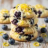 Macro close-up of golden-brown Zesty Lemon Blueberry Cheesecake Cookies with lemon glaze, fresh blueberries, and lemon zest on a rustic wood surface