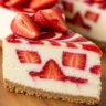 A close-up of a slice of Velvet Strawberry Dream Cheesecake with a graham cracker crust and strawberry swirl, garnished with fresh strawberries and glaze.