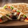 Macro close-up of a sizzling Philly steak and cheese quesadilla cut open, showing gooey melted cheese, beef, and peppers inside a crispy golden tortilla.