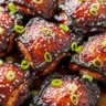 Macro close-up of glossy, sticky Char Siu Chicken with sesame seeds and green onions on a rustic wood board.