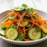 Macro close-up of a vibrant Asian Carrot Cucumber Salad in a rustic bowl, garnished with sesame seeds and cilantro.