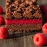 Macro close-up of a decadent dark chocolate raspberry bar slice, showing rich chocolate crust, vibrant raspberry filling, and chocolate crumble topping.
