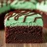 A close-up of a perfectly cut Emerald Isle Mint Fudge Brownie with vibrant green frosting and chocolate drizzle.
