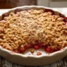 A macro close-up of a golden-brown rhubarb crisp, bubbling with a jewel-toned, jammy rhubarb filling, served in a rustic baking dish on a wooden table.