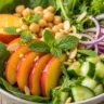 Macro close-up of a vibrant Glow-Up Peach & Chickpea Power Bowl with Zesty Lime Drizzle, featuring sliced peaches, chickpeas, mixed greens, and fresh mint.